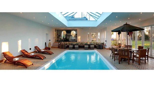 Un'ampia area piscina interna con lucernario, poltrone relax, un tavolo da pranzo e vista sull'esterno.