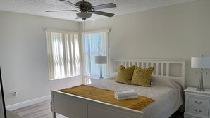 Un dormitorio con cama, ventilador de techo, mesita de noche y ventana con cortinas.