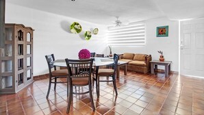 Un coin repas comprenant une table ronde, quatre chaises, un ventilateur de plafond, un canapé en cuir marron et une petite table d’appoint sur laquelle est posé un vase.