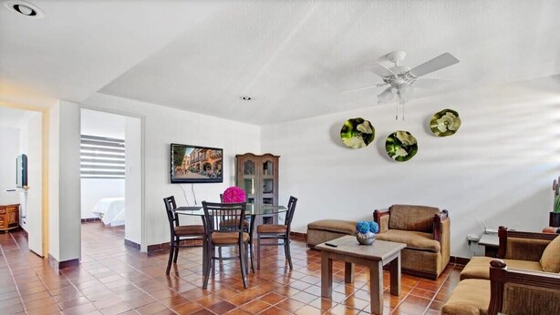 Un vaste espace de vie comprenant une table à manger, des chaises et un canapé. On y trouve également un ventilateur de plafond, un tableau au mur et une fenêtre avec des stores.