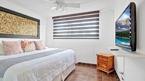 Une chambre à coucher comprenant un lit, un ventilateur de plafond, une fenêtre avec des stores, un téléviseur posé sur un meuble et une petite armoire en bois.