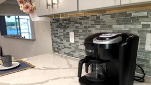 Une cuisine moderne avec une machine à café Keurig posée sur un plan de travail en marbre blanc, une fenêtre donnant sur un plan d’eau et un pare-feu décoré de carreaux gris et blancs.
