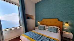 Quarto de hotel com cama, mesa de cabeceira, janela com cortinas e vista para o mar.
