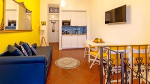 Um espaço compacto com sofá, uma mesa de jantar com cadeiras amarelas, cozinha com armários brancos e uma TV de tela plana fixada na parede.