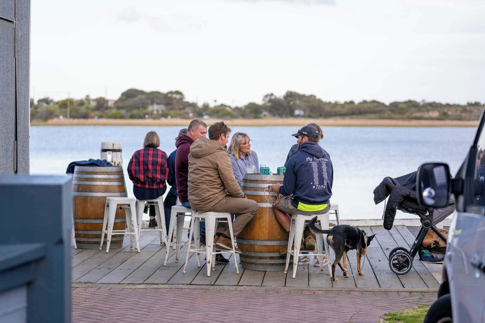 A group of people sitting at wooden tables on a dock by a body of water, with barrels and a stroller nearby.