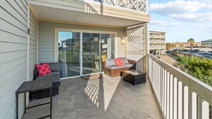 A balcony with wicker furniture, a glass door, and a view of the street. - Beach Condo by 61st Fishing St Pier • Clean, Loved & Steps to Beach, heated pool (Galveston)