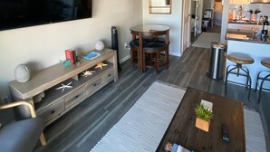 A modern living room with a flat-screen TV, a wooden console table, a dining area with a table and chairs, and a kitchen area with a microwave and stools. - Beach Condo by 61st Fishing St Pier • Clean, Loved & Steps to Beach, heated pool (Galveston)