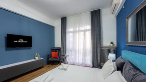 Une chambre d’hôtel moderne avec un grand lit, un téléviseur fixé au mur, une petite table avec une plante et une chaise dotée d’un coussin orange.