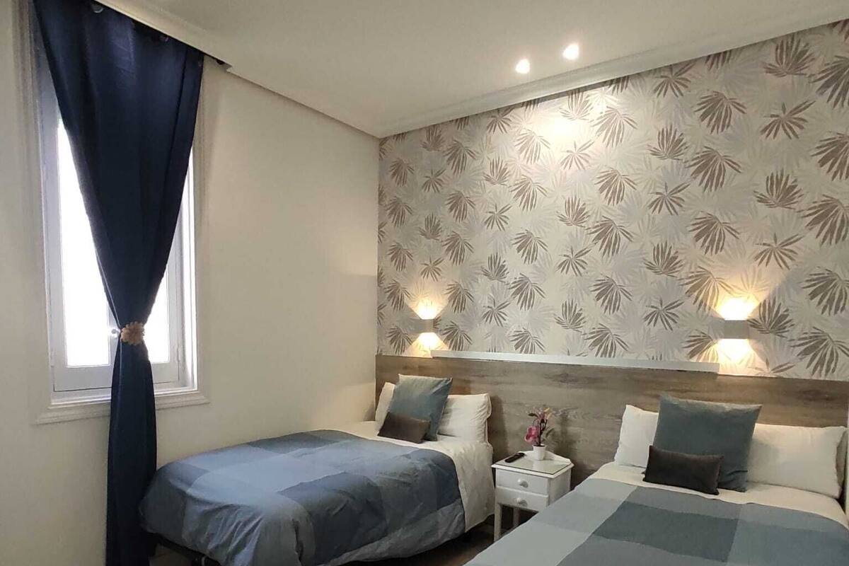 A room with two beds, a chair, a small table, and a window with curtains.