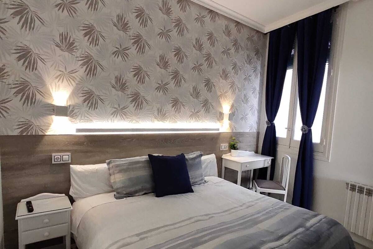 A modern bedroom with a large bed, a bedside table, a nightstand, a small table with a chair, and a window with curtains.