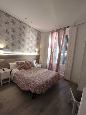 A bedroom with a large bed, a bedside table, a nightstand, a lamp, and a window with curtains. - Hostal Alfaro (Madrid)