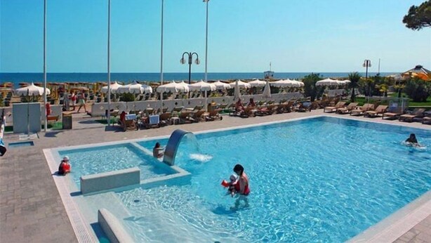 A swimming pool with lounge chairs and umbrellas, flags, and a clear view of the sea. - Cesare Augustus (Jesolo)