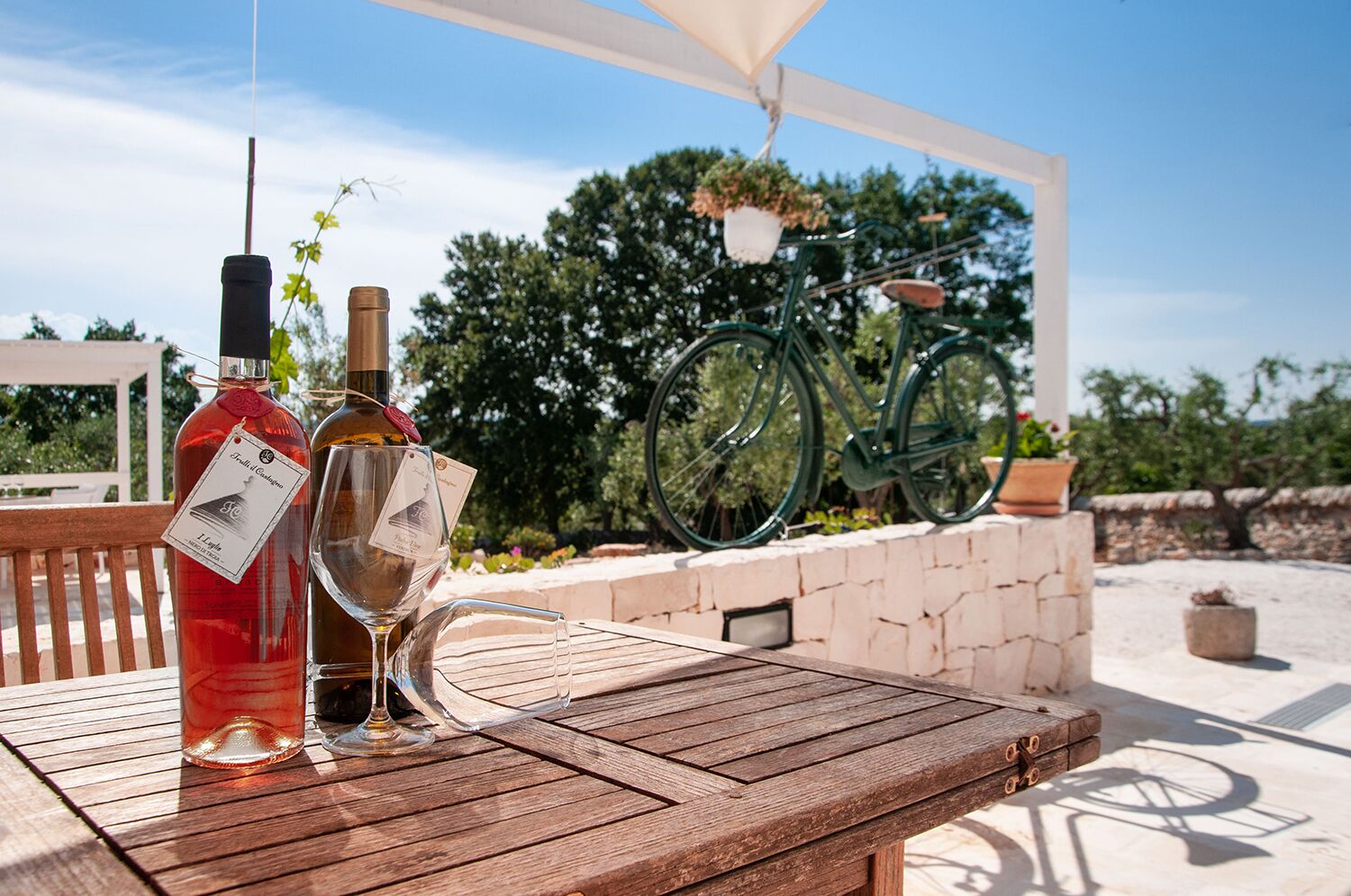 A wooden table with two wine bottles and a wine glass on it, set outdoors with a bicycle and greenery in the background.