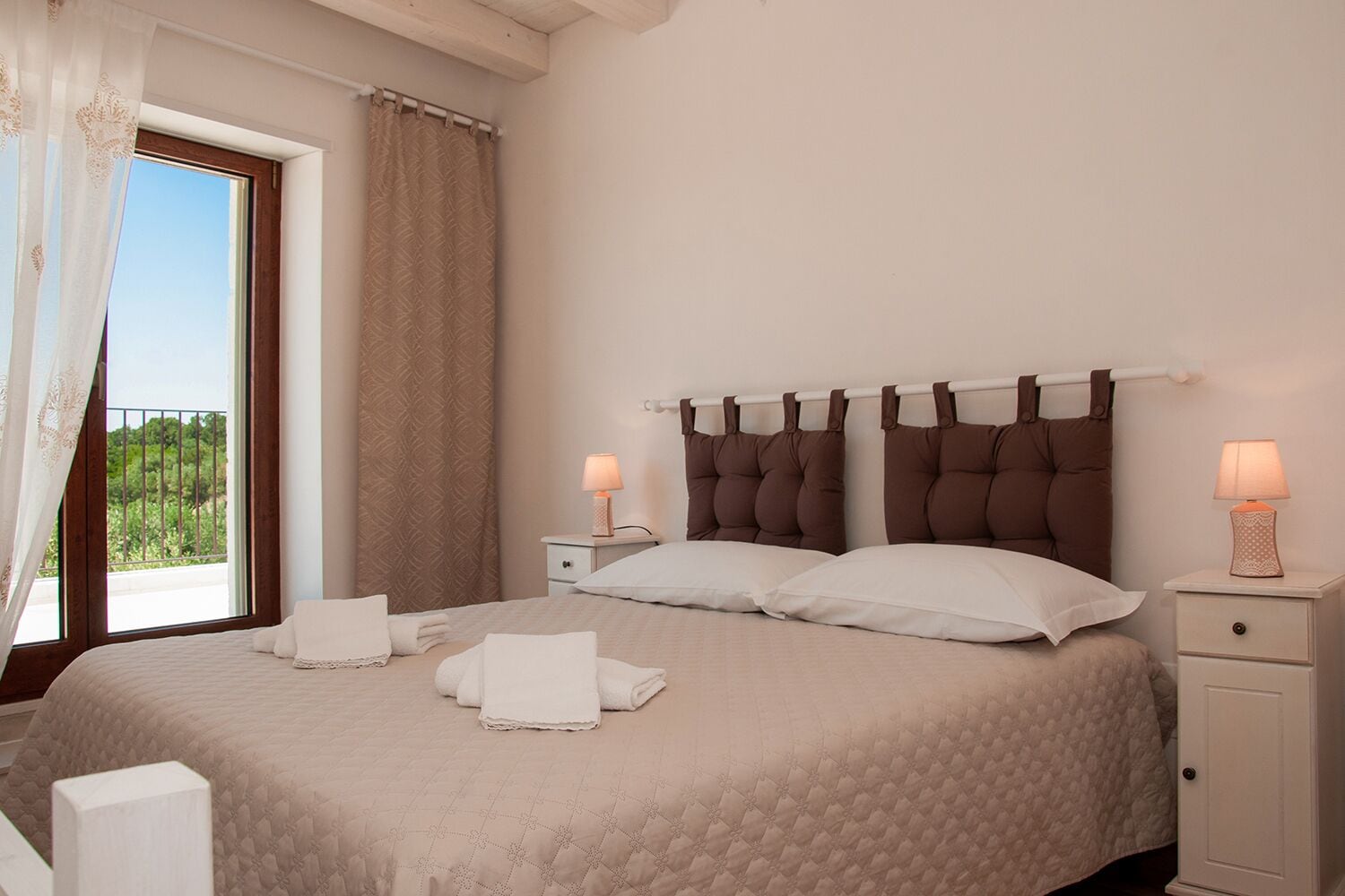 A neatly made bed with white linens, two bedside tables with lamps, and a window with a view of greenery.