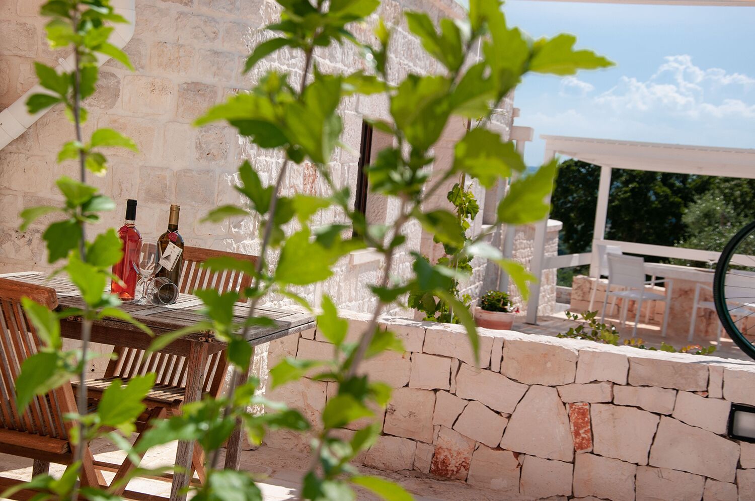 Un espace de restauration en plein air agrémenté d’un mur en pierre, de chaises en bois et d’une table où sont disposées des bouteilles de vin et des verres.