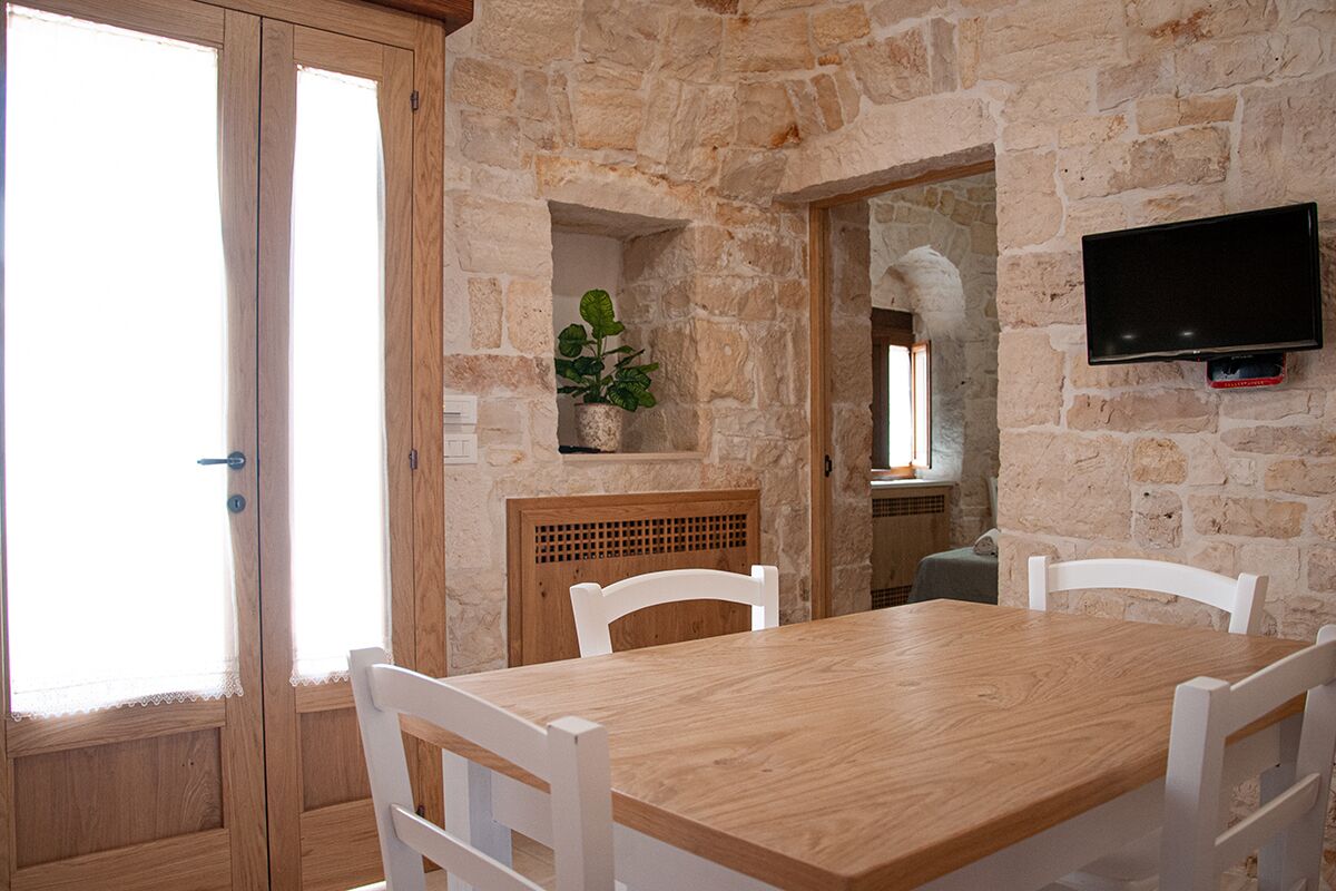 Un coin repas avec une table en bois et des chaises blanches, des murs en pierre, une plante en pot et un téléviseur fixé au mur.