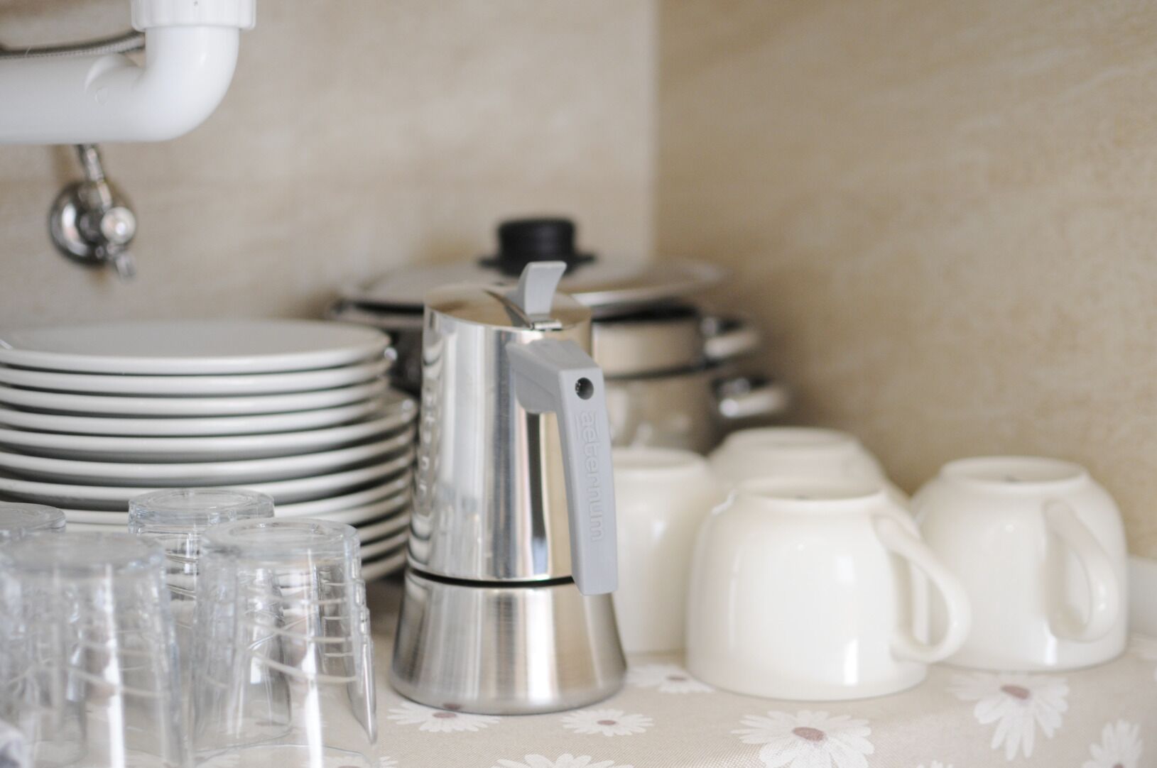 A kitchen area with stacked white plates, a stainless steel coffee maker, and clear glass tumblers.