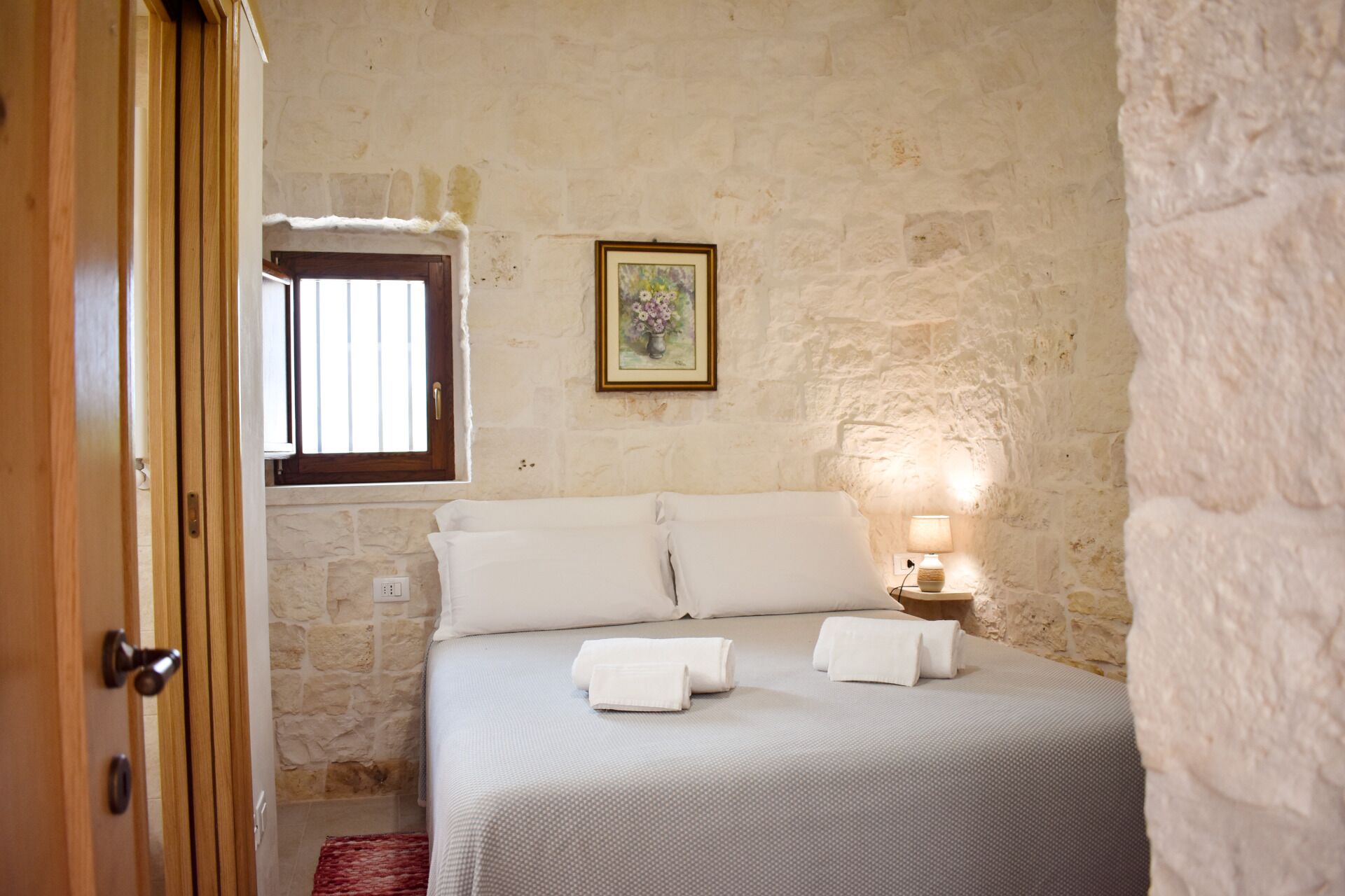 A bedroom with a stone wall, a bed with white linens, a bedside lamp, and a window with bars.