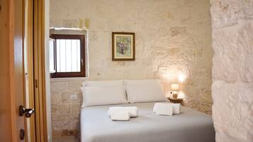 A bedroom with a stone wall, a bed with white linens, a bedside lamp, and a window with bars.