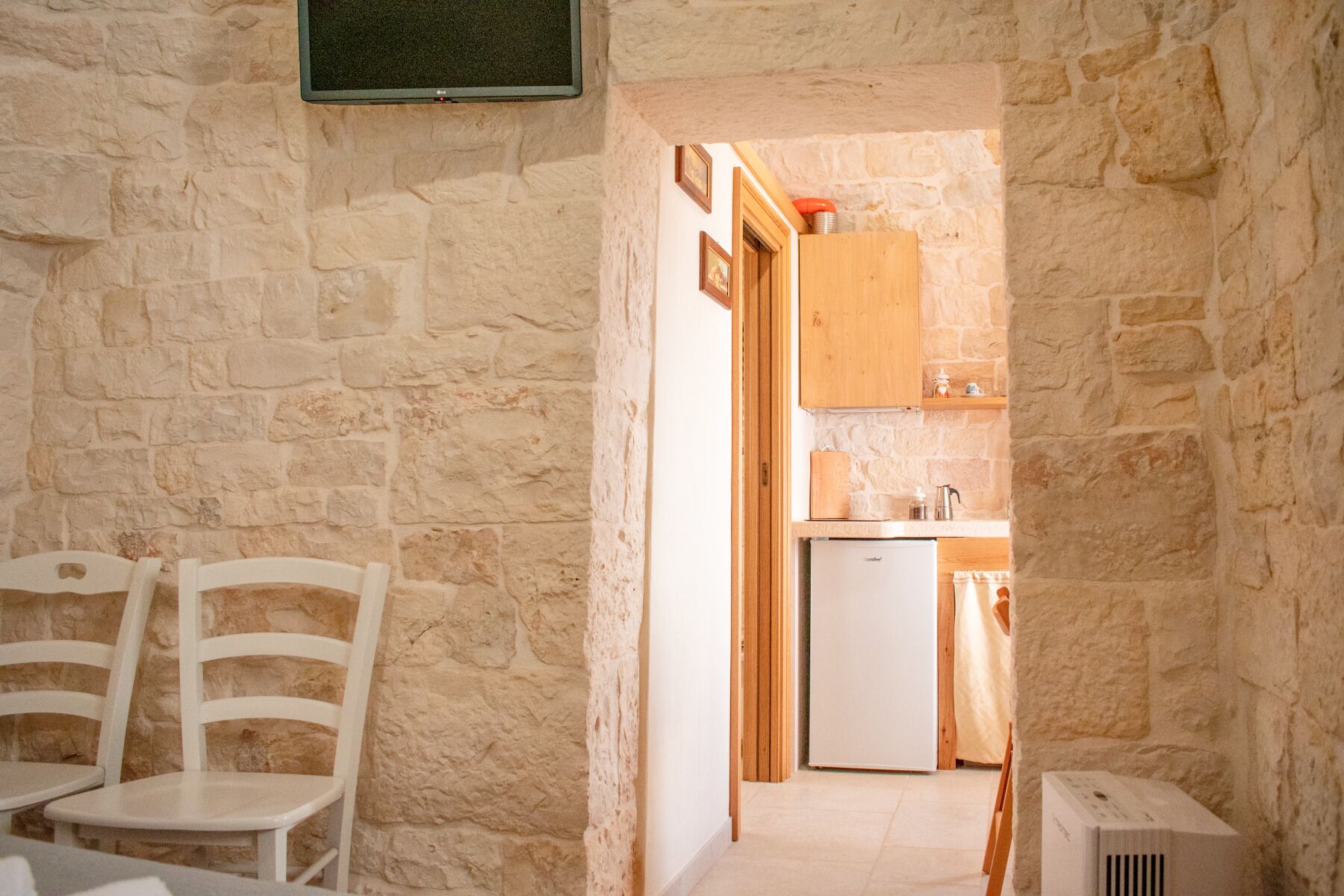 Una habitación estrecha con paredes de piedra, dos sillas blancas y una mesa pequeña. Un acceso da a una zona de cocina con un refrigerador, un armario de madera y un calentador pequeño.
