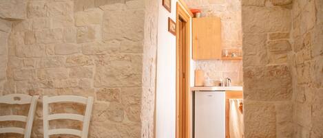 A narrow stone-walled room with two white chairs and a small table. A doorway leads to a kitchen area with a refrigerator, a wooden cabinet, and a small heater.