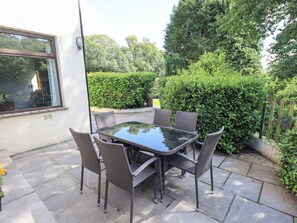 An outdoor patio with a glass-top table and wicker chairs, surrounded by stone tiles and greenery. - Greenbank Cottage (Kendal)