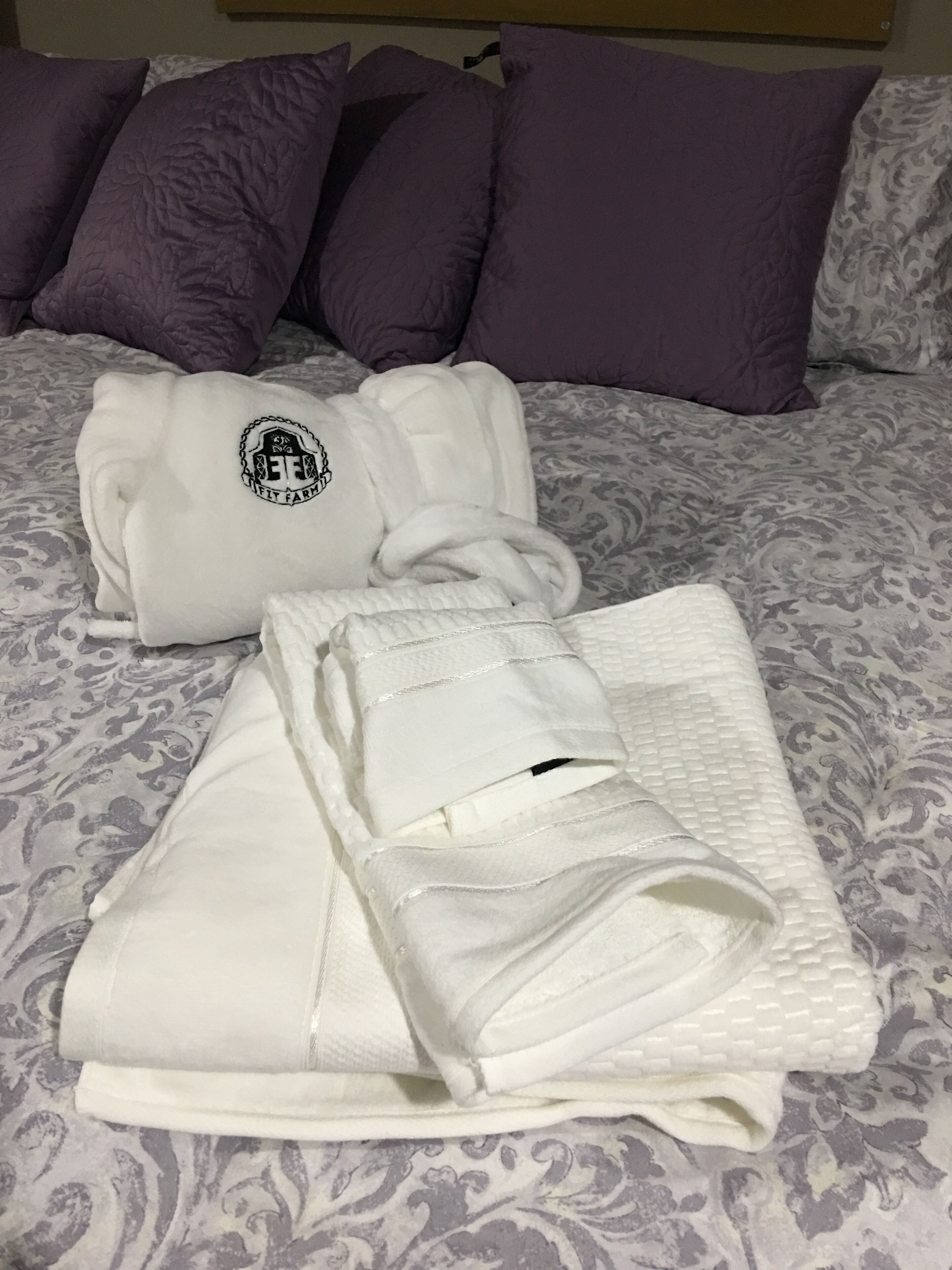 A neatly made bed with a patterned bedspread, a white towel, and a white robe with a logo.