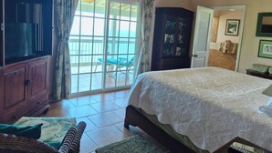 A bedroom with a large bed, a wardrobe, a television, and a view through a sliding door to an outdoor area with chairs. - Nancy's Folly Villa - Fabulous Home - overlooking dazzling Rendezvous Bay (Cruz Bay)
