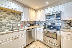 Features a sleek kitchen with stainless steel appliances, microwave, oven, and dishwasher, set against a mosaic tile backsplash.