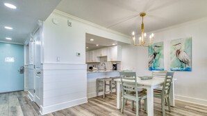 A modern kitchen with white cabinetry, a dining area with a table and chairs, and two wall-mounted paintings of birds. - B105 | Beachfront with Beach Service, Pool, & WiFi (Miramar Beach)