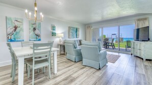 A spacious living area with a dining table and chairs, a sofa, a chandelier, artwork, and a large window with a view. - B105 | Beachfront with Beach Service, Pool, & WiFi (Miramar Beach)