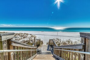 Una escalera de madera que lleva a una playa, con cielo azul despejado y vista al océano.
