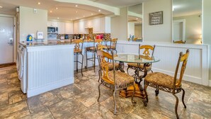 A kitchen with a central island, white cabinetry, and a dining area with ornate chairs. - C103 | Beachfront with Beach Service, Pool, & WiFi (Miramar Beach)