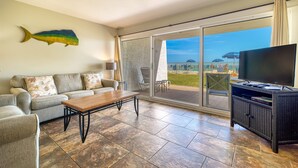A living room with a sofa, coffee table, TV stand, and a large sliding glass door leading to a balcony with a view of the sea. - C103 | Beachfront with Beach Service, Pool, & WiFi (Miramar Beach)