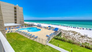 Uma área de piscina com cadeiras e guarda-sóis azuis, uma praia com areia branca e um céu azul e límpido.