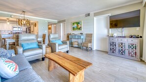 Uma sala de estar ampla com sofá, poltronas e uma mesa de centro de madeira. Há uma televisão fixada na parede, e uma área de cozinha visível ao fundo.