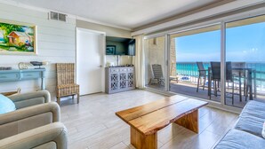 Uma sala de estar ampla com uma mesa de centro de madeira, um sofá e uma televisão fixada na parede. Há uma varanda com vista para o oceano.