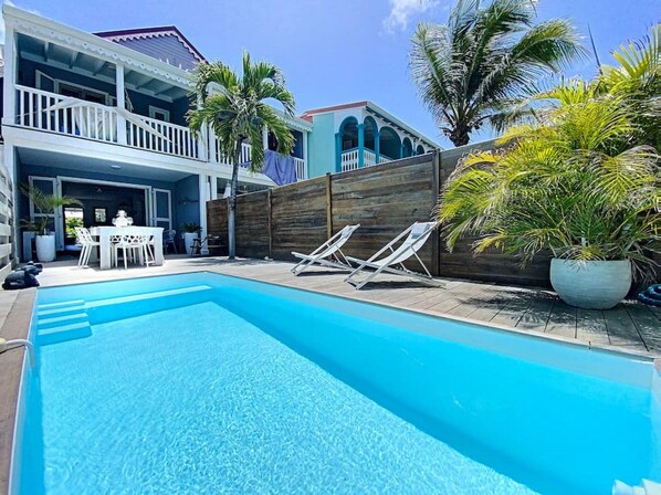 A swimming pool with lounge chairs, a dining area with a table and chairs, and a wooden fence. - Villa Casalilo, piscine privée, à 100m de la plage d`Orient Bay, 2 chambres (Orient Bay)