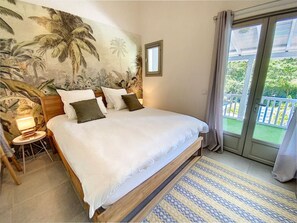 A bedroom with a large bed, a nightstand, a lamp, a mural of palm trees, and a balcony with a view of greenery. - Villa Casalilo, piscine privée, à 100m de la plage d`Orient Bay, 2 chambres (Orient Bay)