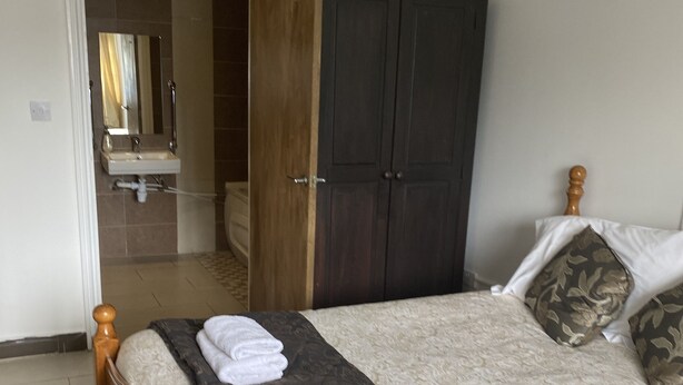 Een net opgemaakt bed met een bedspreide met patroon en twee opgerolde handdoeken, een donker houten kledingkast, en een badkamer zichtbaar door een geopende deur.