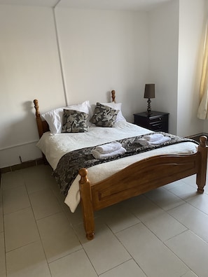 A neatly made bed with a wooden headboard, a dark patterned bedspread, and several pillows, including floral ones. There is a small bedside table with a lamp and a dark-colored nightstand. The room has white walls and a tiled floor. - 2 En-suite Bedrooms Flat in Kidlington ,oxford (Kidlington)