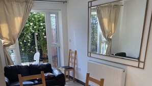 Een woonkamer met een zwarte leren bank, een houten eettafel met vier stoelen, een radiator en een grote spiegel aan de muur.