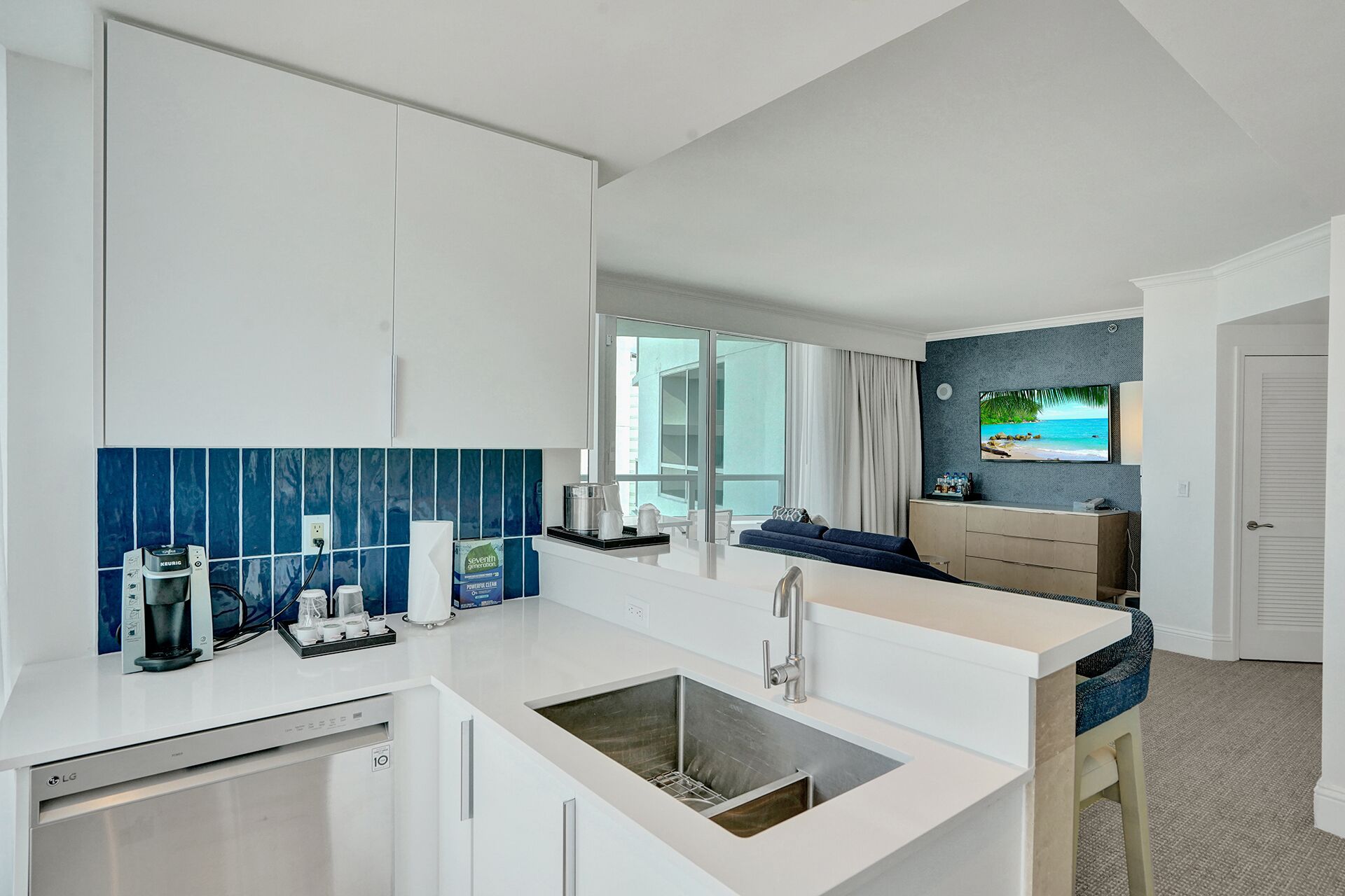 A modern kitchen with white cabinets, a built-in sink, and a coffee maker. There is a large window with a view of the ocean and a TV mounted on the wall.