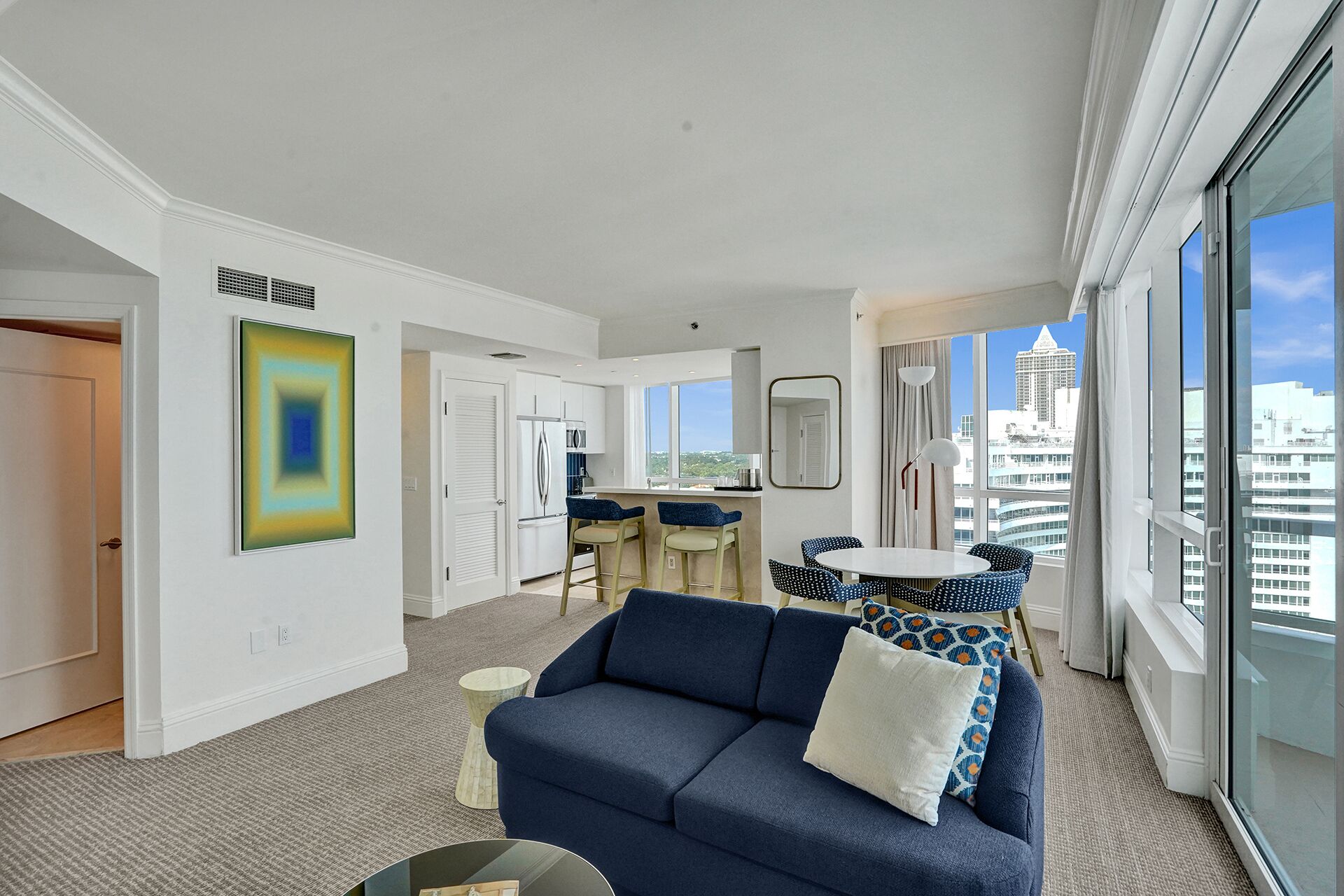 A modern living room with a blue sofa, a dining table with chairs, a kitchen area, and large windows offering a city view.