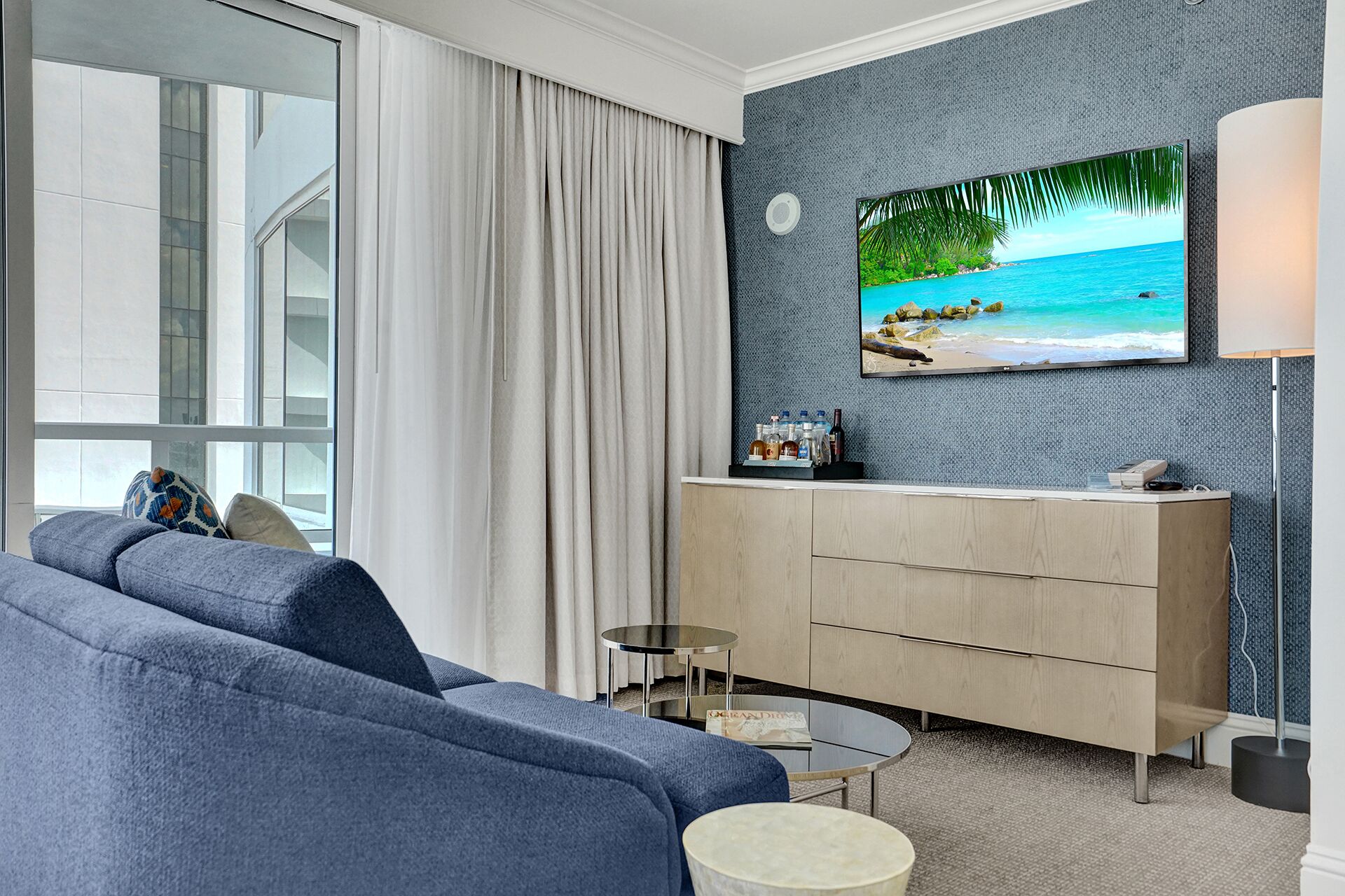 Habitació d'hotel moderna amb una finestra gran, un sofà gris, un armari de fusta, una taula rodona petita i un televisor fixat a la paret que mostra una escena de platja.