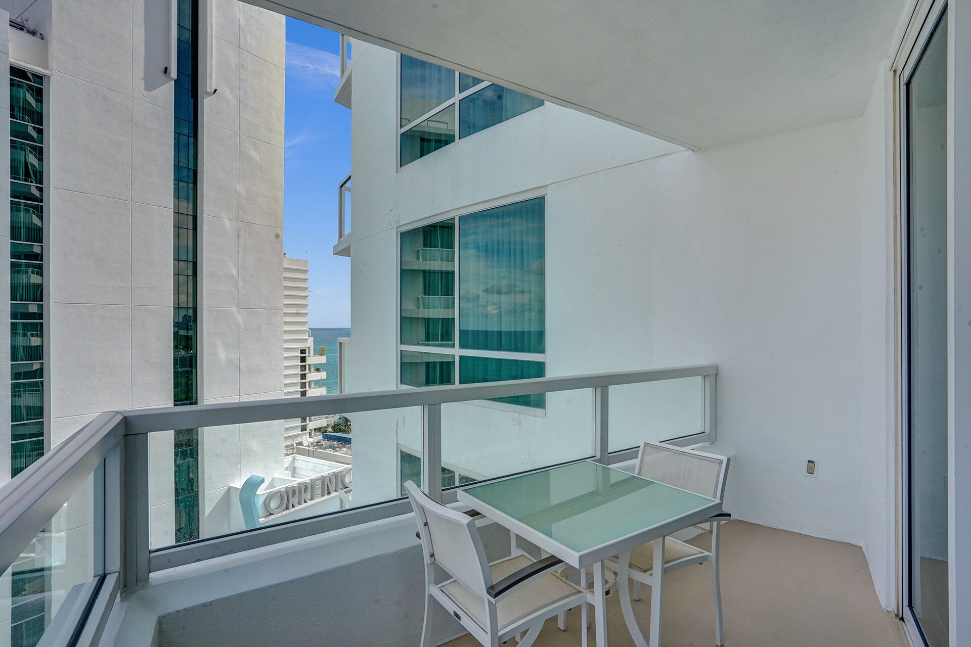Varanda com mesa e cadeiras de vidro, com vista para um edifício moderno com grandes janelas.