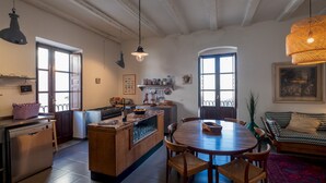 A spacious kitchen with a central island, wooden dining table, and chairs. There are two windows providing natural light, and a hanging light fixture above the island.