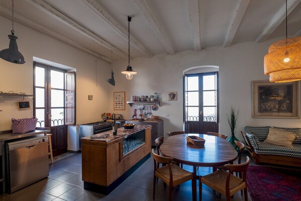 Une cuisine spacieuse dotée d’un îlot central, d’une table à manger en bois et de chaises. Deux fenêtres laissent entrer la lumière naturelle, et un luminaire suspendu est placé au-dessus de l’îlot.