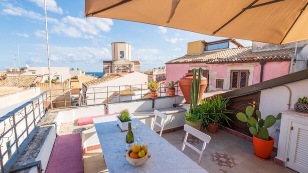 A rooftop terrace with a table, chairs, potted plants, and a view of the cityscape.