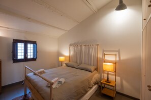 Une chambre à coucher avec un lit, une table de chevet, une lampe, une fenêtre avec des rideaux et une chaise en bois.